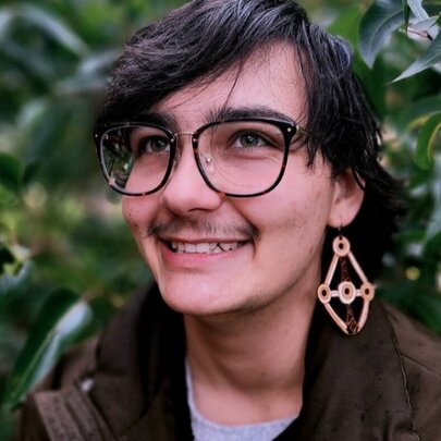 Smiling person with a large earring hanging from their earlobe, wearing glasses on their face, with greenery behind them.
