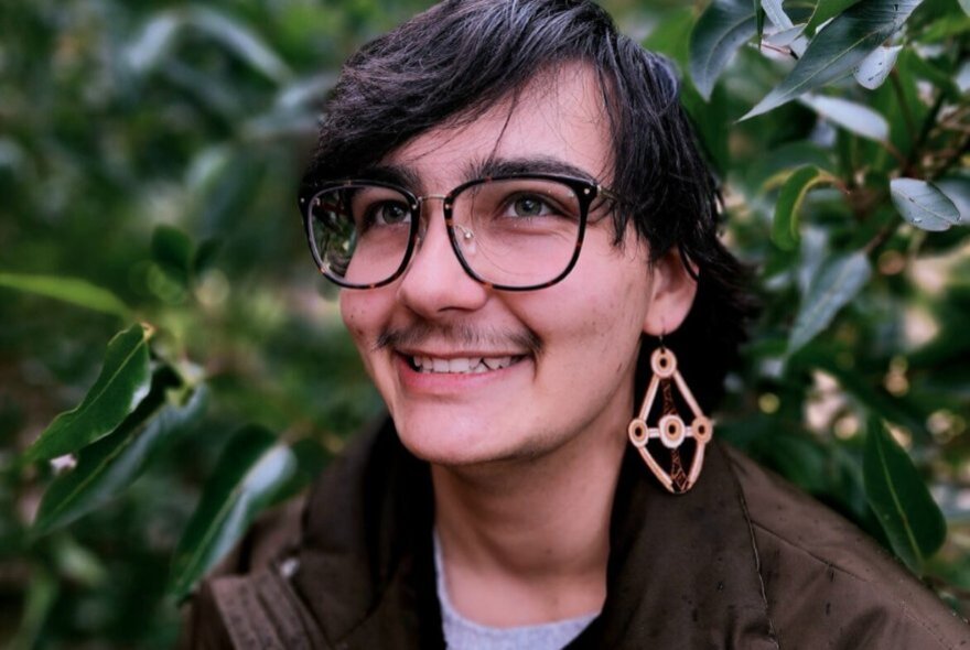 Smiling person with a large earring hanging from their earlobe, wearing glasses on their face, with greenery behind them.