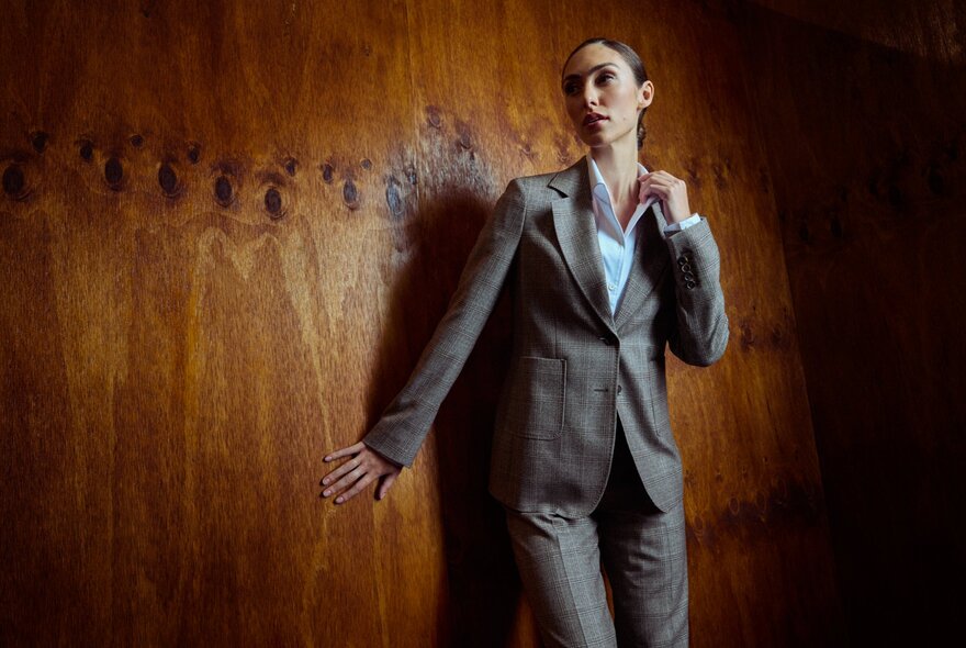 A female model wearing a tailored pants suit with an open collar shirt, leaning against a wooden wall.