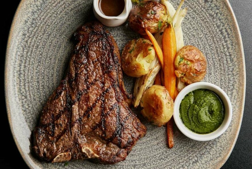 A pottery plate of food featuring a large steak served with baked vegetables and sauces.