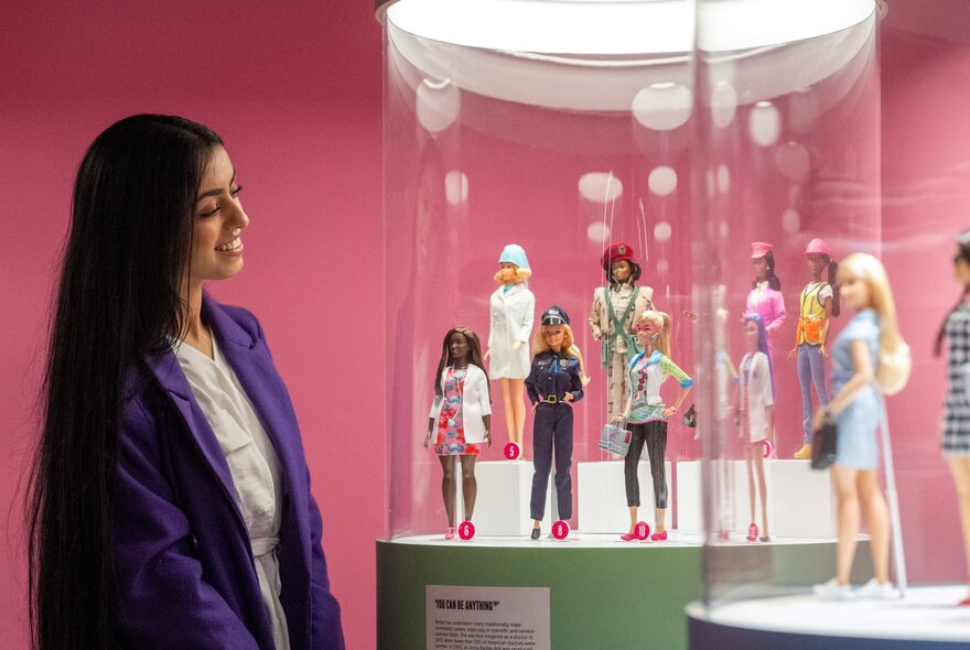 A woman with long dark hair wearing a purple blazer looks at a museum display case filled with various Barbie dolls against a pink background.