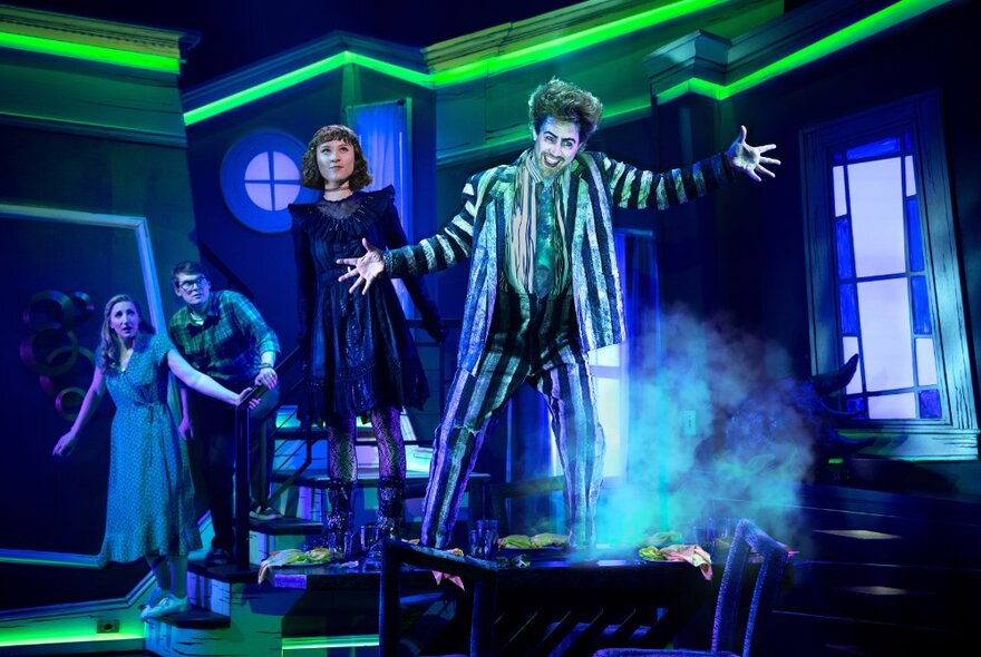Beetlejuice lead actor Eddie Perfect in costume with green hair and striped grey suit, smiling with arms outstretched, on stage with other cast members.