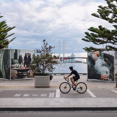 Large scale artworks depicting smoking ceremonies, outdoors at Melbourne's Docklands, a lone cyclist riding past. 