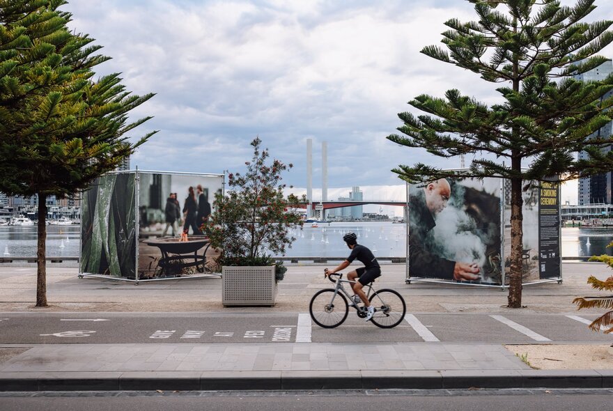 Large scale artworks depicting smoking ceremonies, outdoors at Melbourne's Docklands, a lone cyclist riding past. 