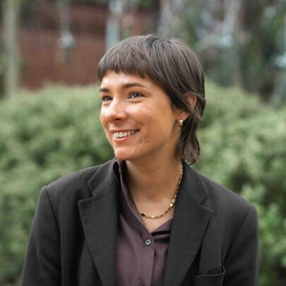 Goorie and Koori poet, Dr Evelyn Araluen, smiling for a portrait as she looks off camera, green bushes behind her.