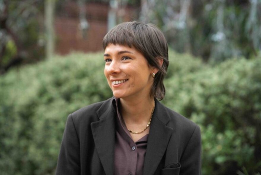Goorie and Koori poet, Dr Evelyn Araluen, smiling for a portrait as she looks off camera, green bushes behind her.