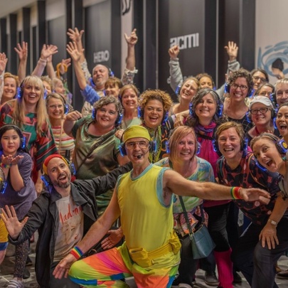 A group of of brightly coloured dancers looking happy, led by man in bright green and yellow shiny outfit.