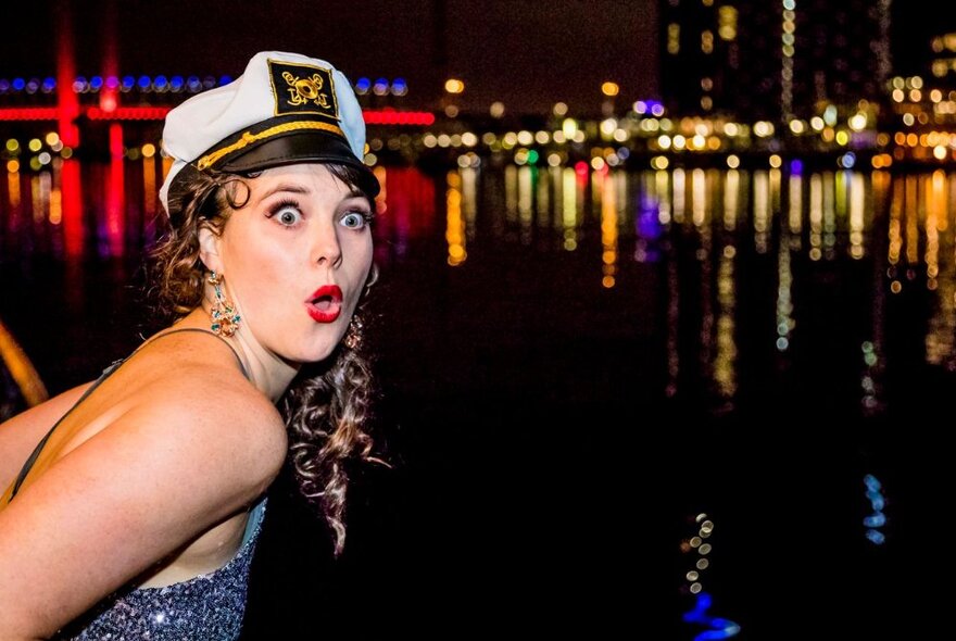 Person wearing a sailor cap, mouthing ooh, with twinkling lights on the water in the background.