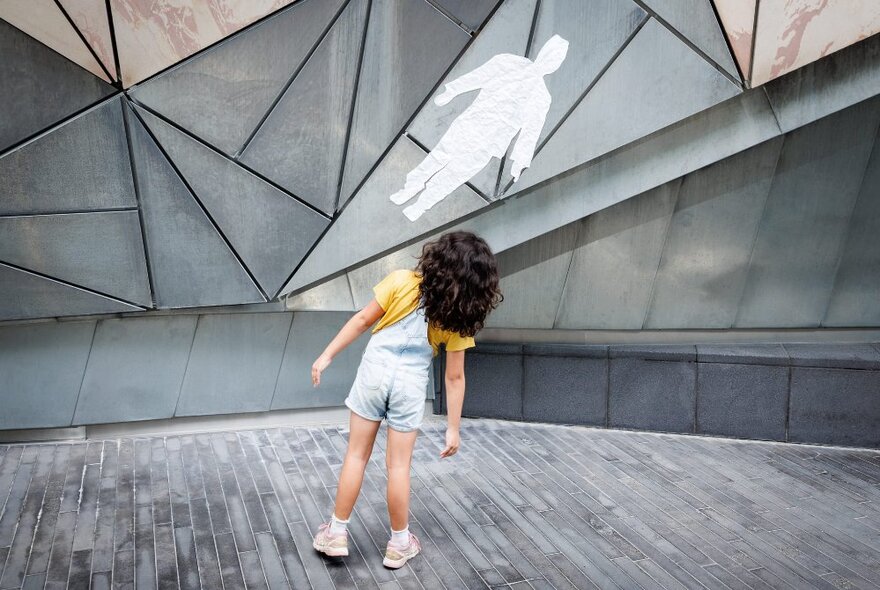 A child wearing overalls looking at a white shape of a person stuck to a wall at Fed Square.