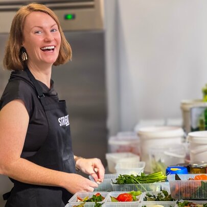 A woman in a black apron laughing as she prepares food at a bench top. 