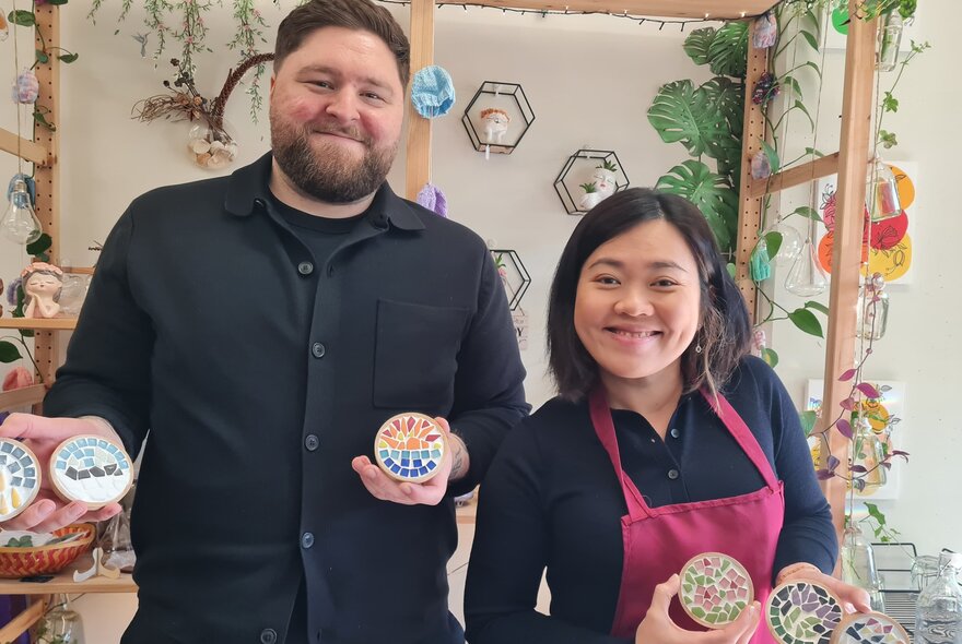 Workshop participants smiling and holding multicoloured mosaic tiled coasters.