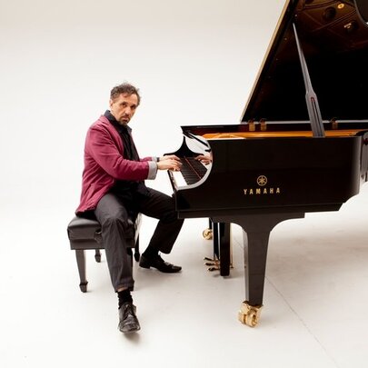 Pianist Joe Chindamo playing a glossy black grand piano against a white background.