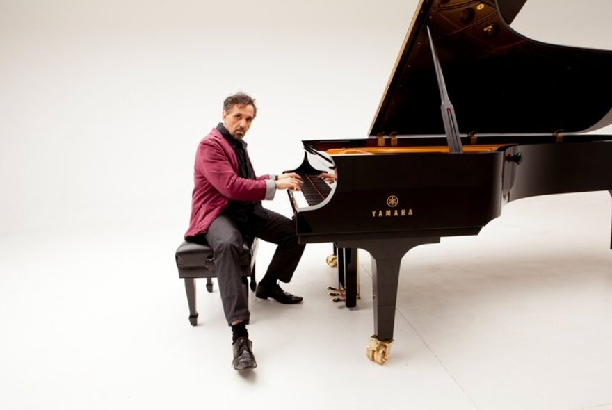 Pianist Joe Chindamo playing a glossy black grand piano against a white background.