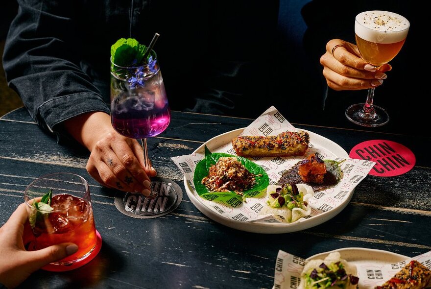 Three sets of hands, each holding up drinks over a dark table, with a plate of Asian snack food in front of them.