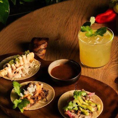 Three tacos served on a wooden platter, with a dipping sauce and a margarita cocktail in a short glass.