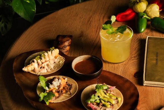 Three tacos served on a wooden platter, with a dipping sauce and a margarita cocktail in a short glass.