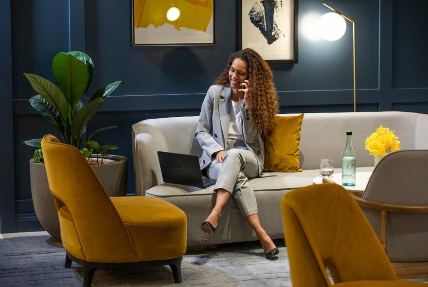 Person in a business suit talking on a mobile phone while sitting on a couch in a lounge area of a hotel, with plants, artwork and soft lighting.