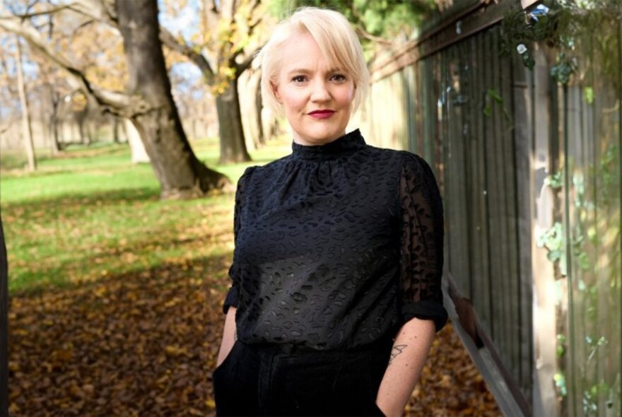 Jacinta Parsons with hands in her pockets standing casually in front of a fence with trees and autumn leaves behind her. 