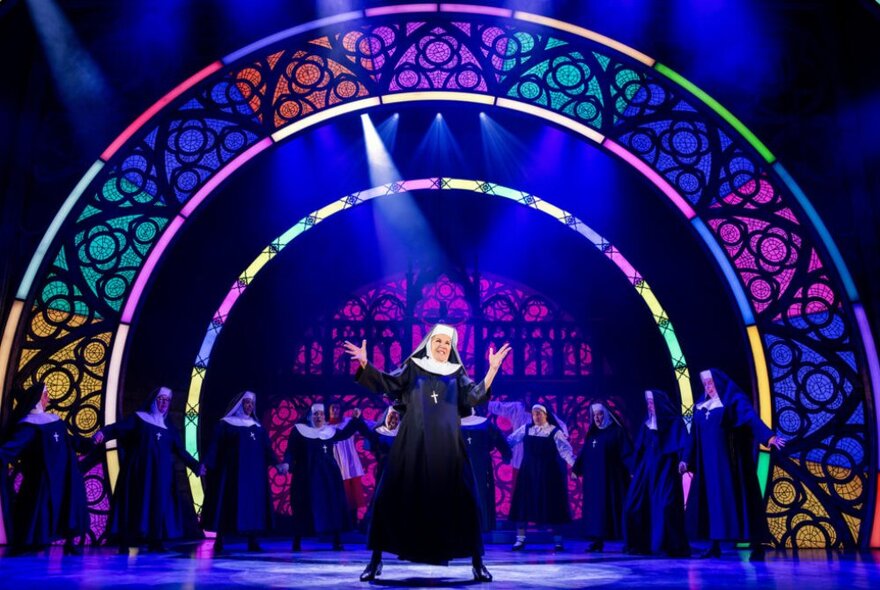 A nun in a black and white habit, singing on stage with a large purple stained glass set decoration behind her.