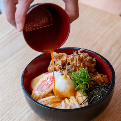 A person is pouring sauce over a bowl of udon noodles with beef, spring onions, seaweed, radish and fish cake