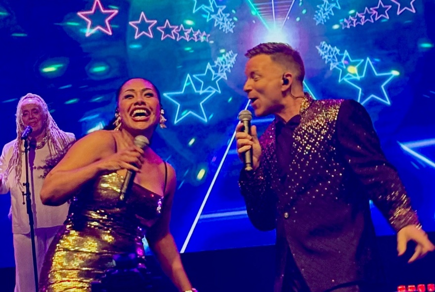 Singers Paulini and Tim Campbell on stage with a back-up singer and a projection of bright neon stars behind them.