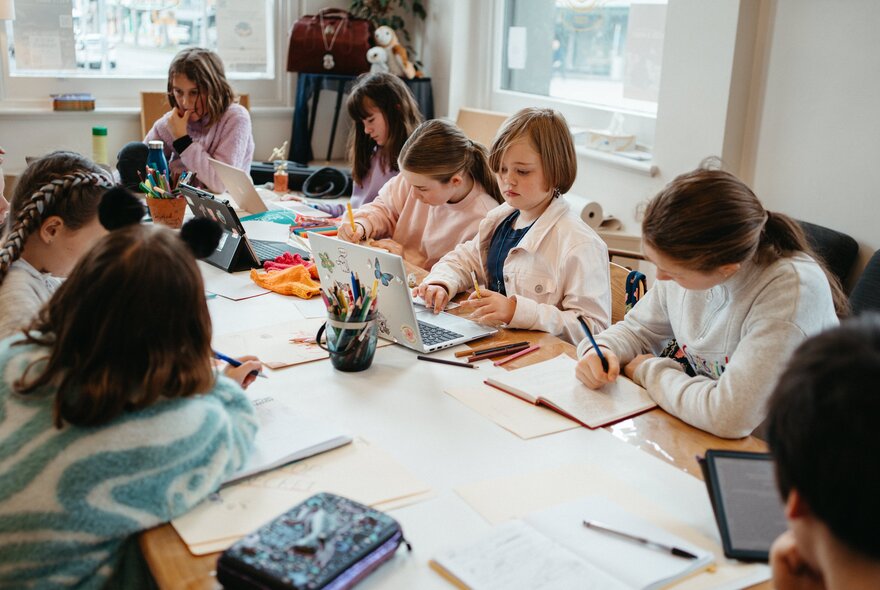Children seated at a large table working with pens, paper and laptops.