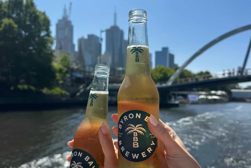 Two hands holding up two small bottles of beer, with a view of the Yarra River and Melbourne city in the background.