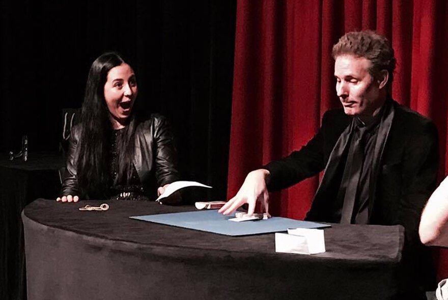 Magician Tim Ellis performing a close-up illusion at a table in front of a red curtain, with an astounded audience member watching with her mouth open in surprise.