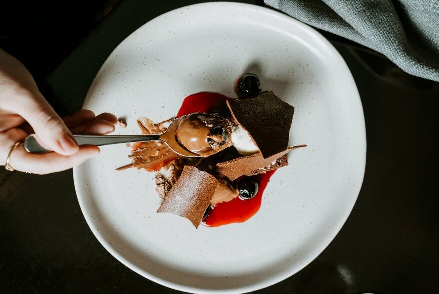 A hand holding a spoon of ice-cream over a plate of a dessert.