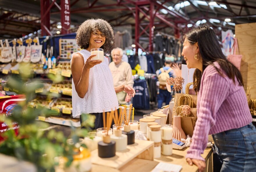 A woman holding up a candle and smiling at a stall holder at a market. 