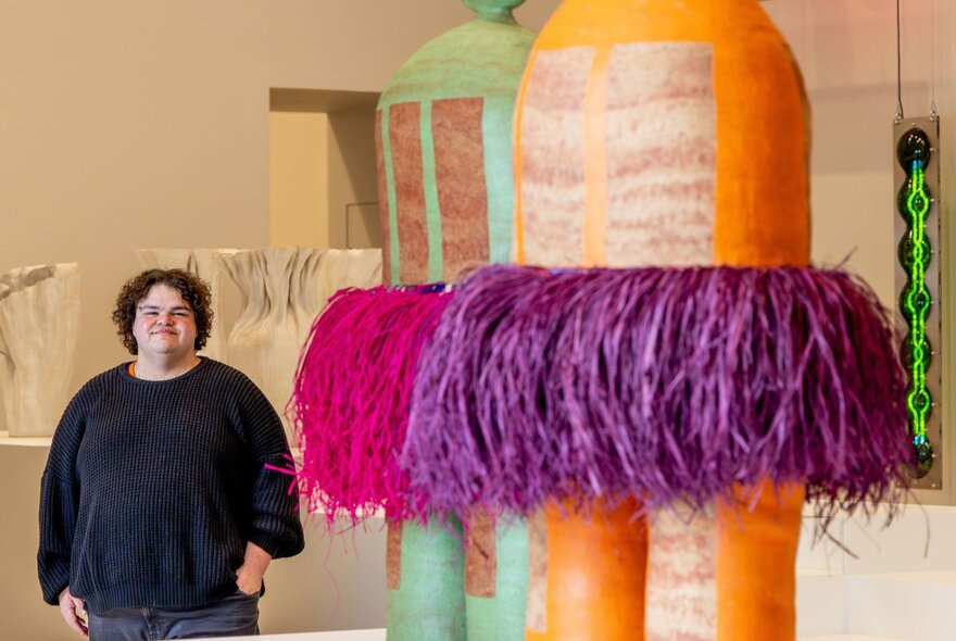 A smiling person standing in front of a large orange and purple design object on display at NGV.