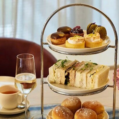A three-tiered cake stand showcasing a selection of small delicacies, alongside a glass of sparkling wine, a teacup and saucer and a small floral arrangement.