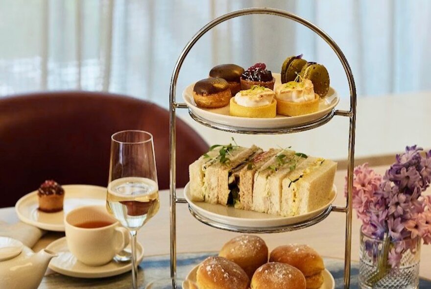 A three-tiered cake stand showcasing a selection of small delicacies, alongside a glass of sparkling wine, a teacup and saucer and a small floral arrangement.