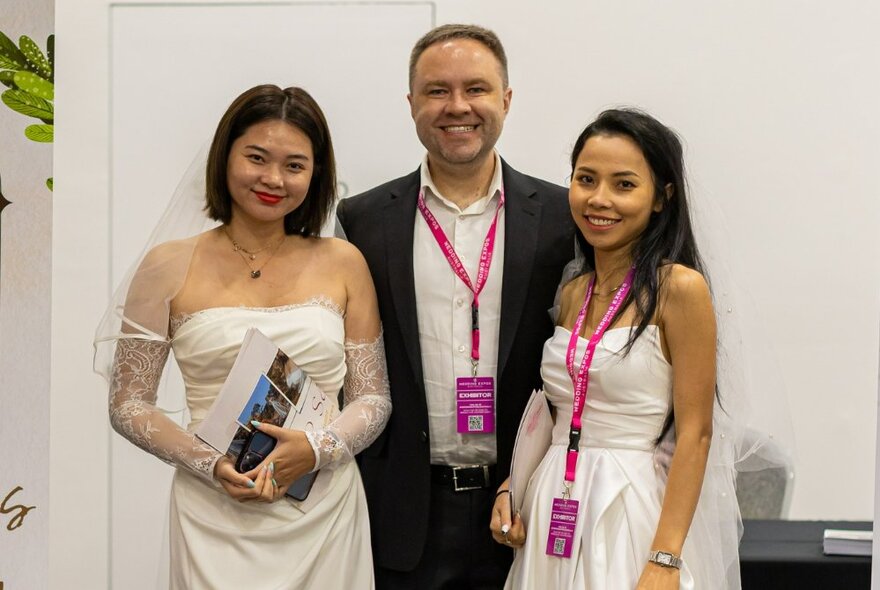 Dark-suited man standing next to women wearing white gowns.