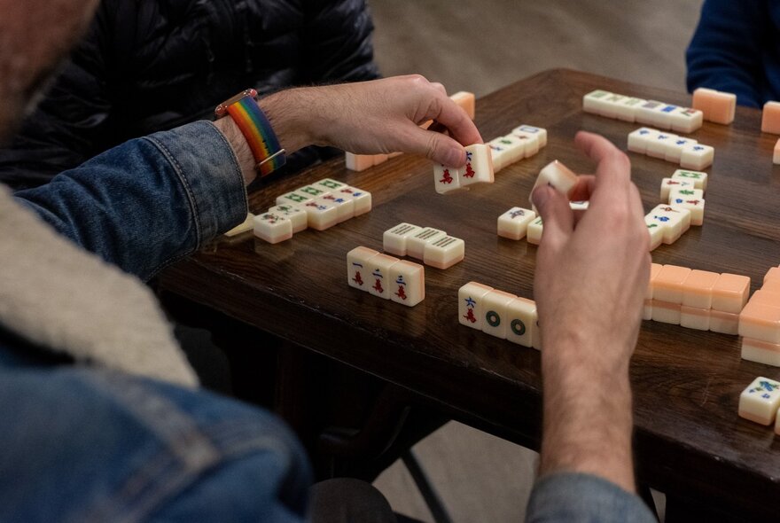 Looking over someone's shoulder as they play mahjong at a wooden table with mahjong pieces on it.