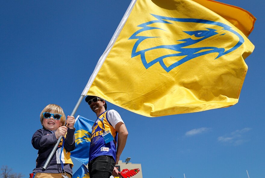 A young football fan waving a West Coast Eagles AFL flag, with a blue sky background.