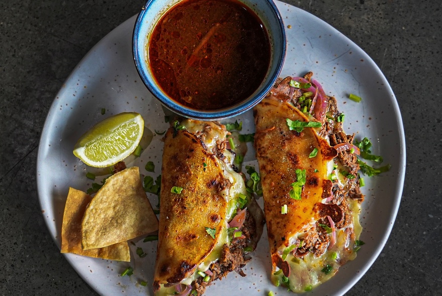 Two massively overstuffed soft tacos on a plate with a lime wedge and a dipping salsa.