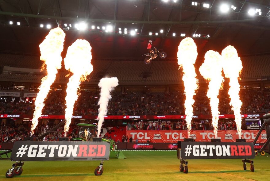 Jest of flames shooting into the air on a sports field, with cricket fans in the stadium seats in the background.