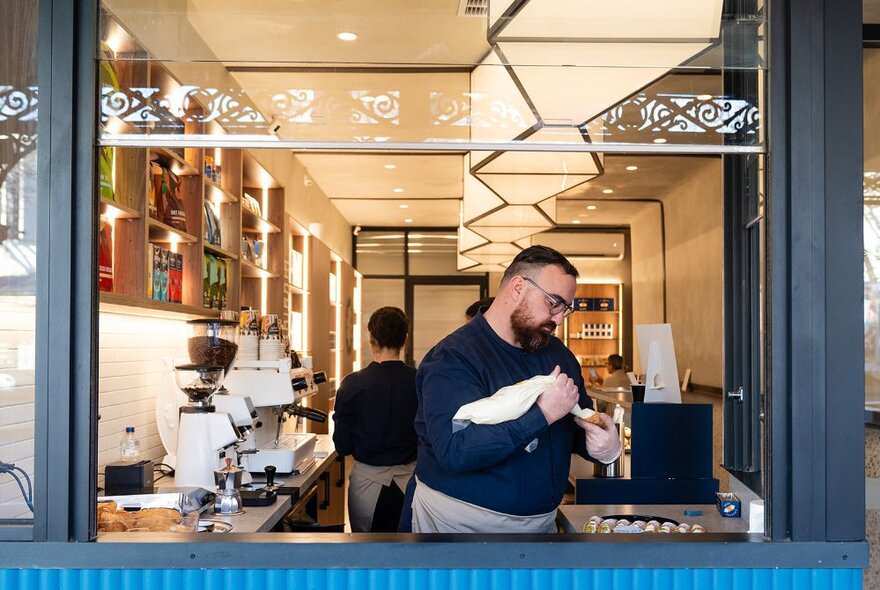 Chef squeezing ricotta into cannoli shells, in the window of a cafe wtih counters and shelves.