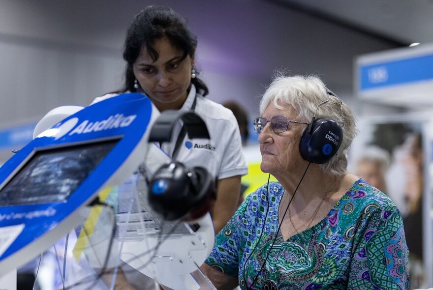 A older person wearing headphones seated at an audio station at an expo, with a person standing alongside looking intently at the display.