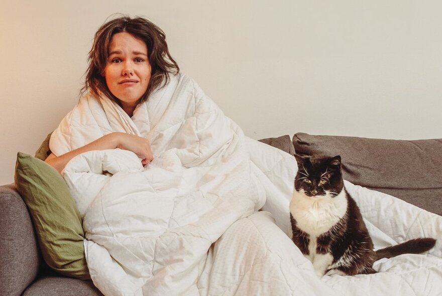 Comedian, Jenna Suffern, seated on a grey couch wrapped in a white doona, with a grumpy looking black and white cat seated next to her.