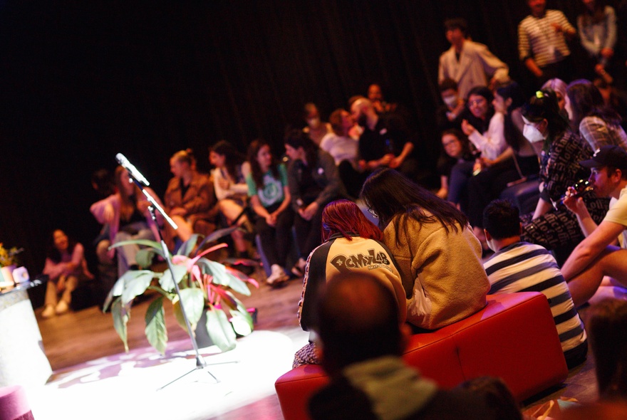 Angled view of people in a theatre setting listening to a lecture, a plant on a table.
