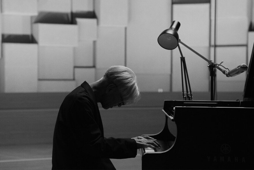 Musician Ryuichi Sakamoto seated at a piano, his hands on the keys and his head bent, lit by an anglepoise lamp.