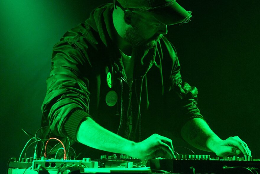 A person wearing a hat and hoodie, illuminated by green light, operates a music mixer or controller.