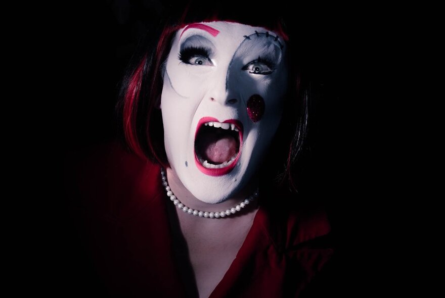 A woman screaming wearing clownish white make-up and pearls.