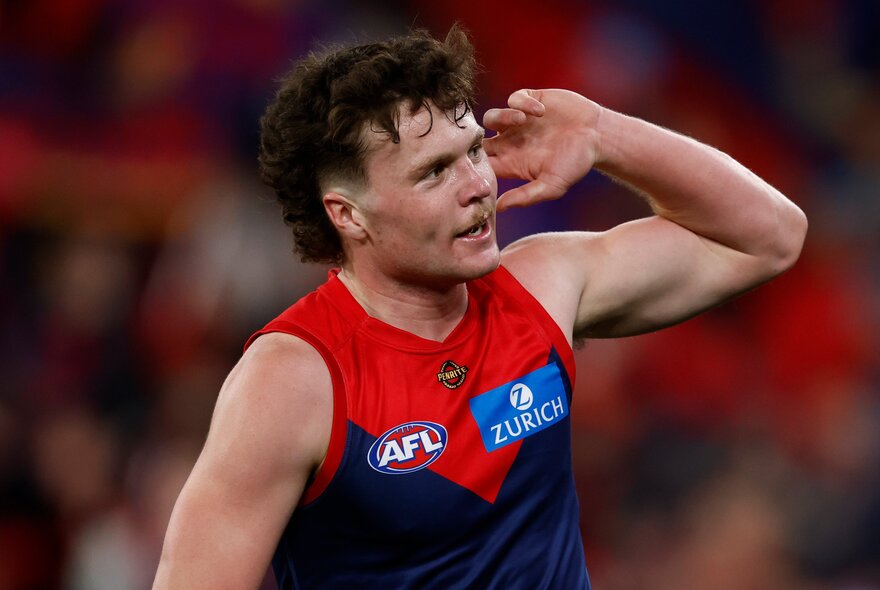 A Melbourne Demons AFL player holding his hand to his ear during a game.