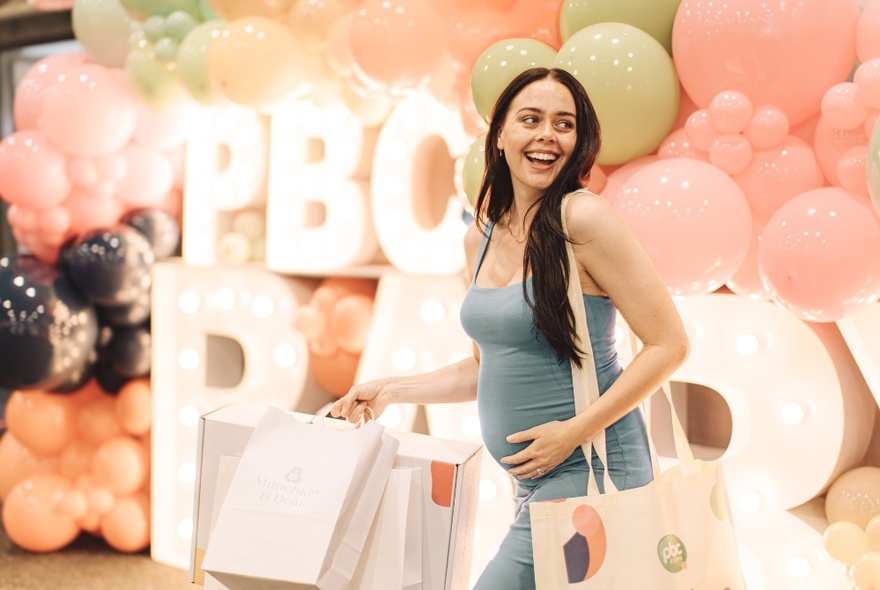 A smiling pregnant woman holding multiple carrier bags at an expo, walking past a large display of balloons.