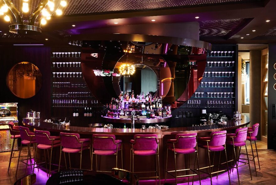 Interior of Dive Bar, showing a circular bar, with high pink velvet bar stools, a wooden floor and large circular mirrors on the rear wall.