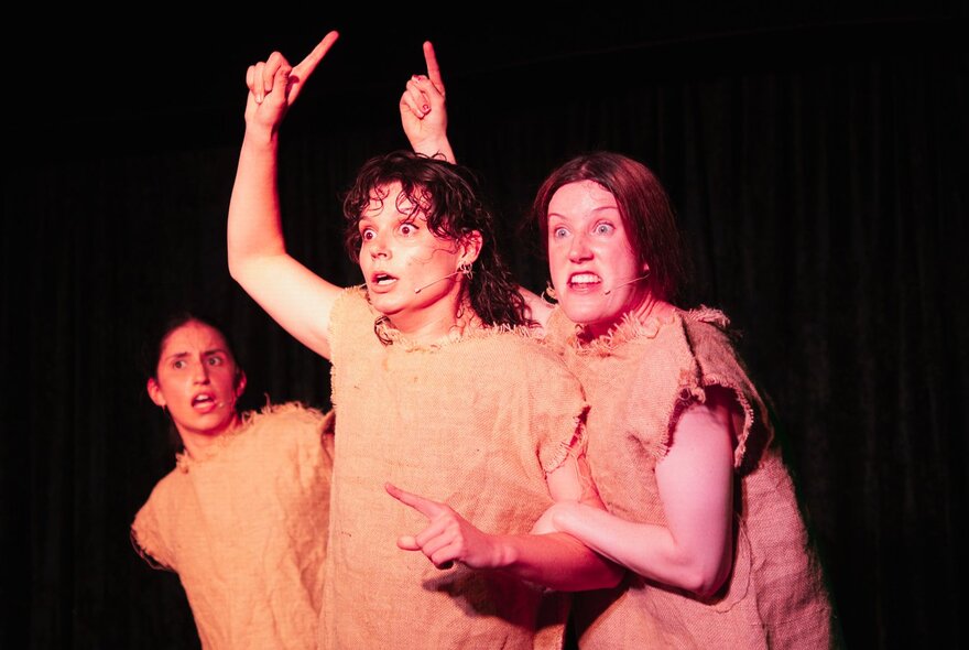 Three women on stage during a performance making exaggerated faces and two holding up single index fingers. 
