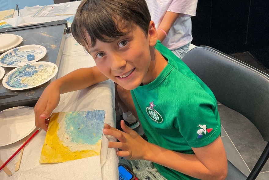 A smiling child turns their head away from a painting they are working on.